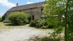Une maison de campagne en pierre, avec une allée gravillonnée, entourée d’une végétation luxuriante et d’arbres.