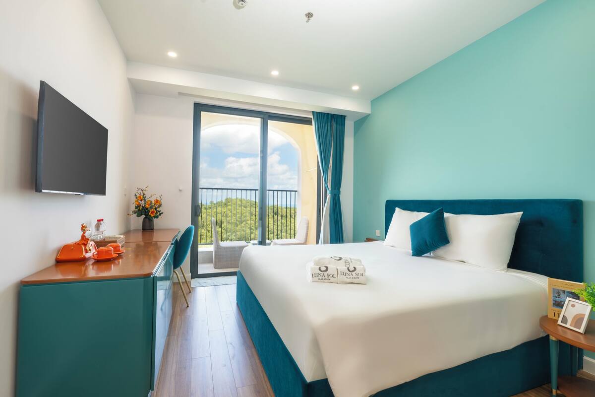 A modern hotel room with a large bed, a desk with a flat-screen TV, and a balcony with a view of greenery.