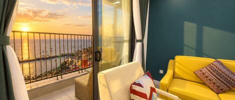 A balcony with a view of the sunset over the sea, a yellow sofa, and a white chair with a patterned cushion.