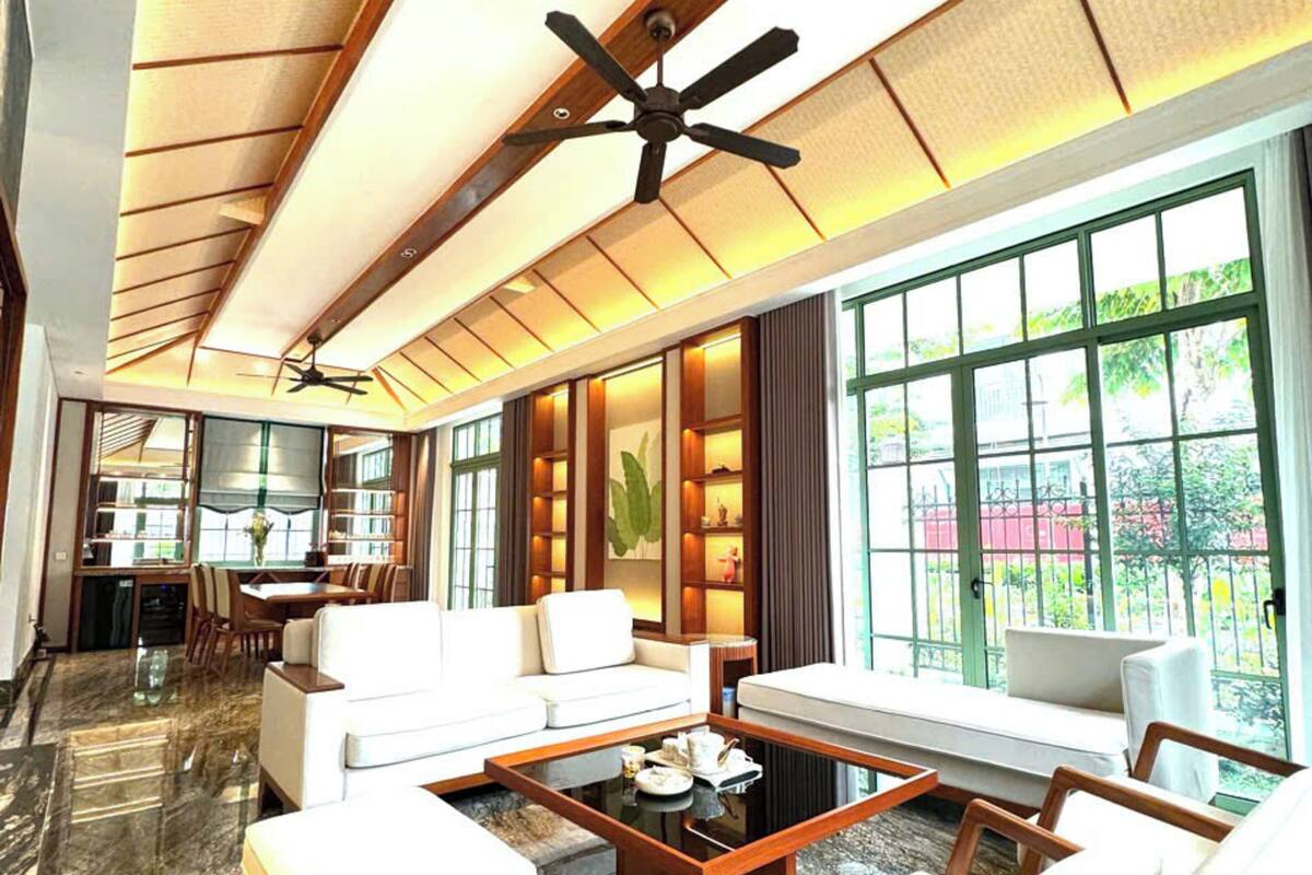 A spacious living room with a high ceiling, a ceiling fan, a dining area with a table and chairs, and large windows offering a view of the outdoors.