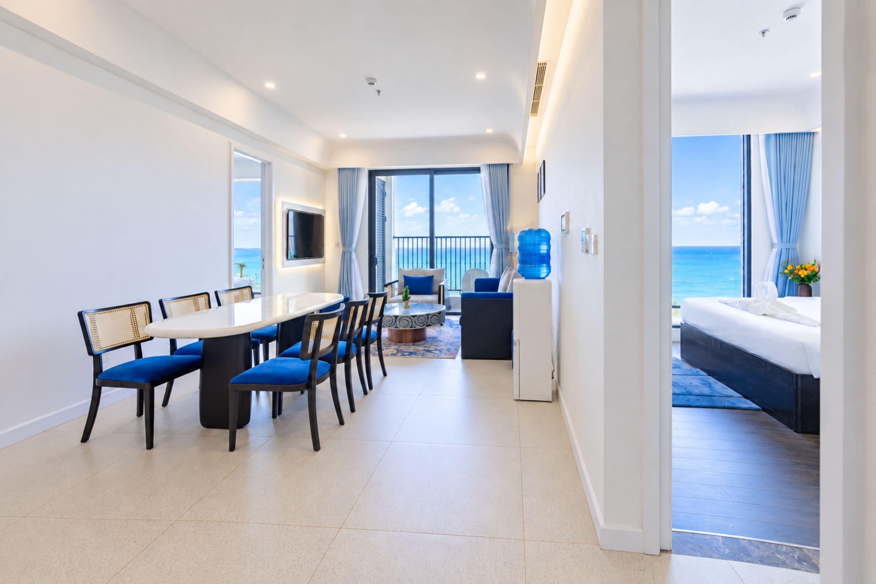 A modern hotel room with a dining area, a sofa, a large window with a view of the sea, and a bed with a view of the ocean.
