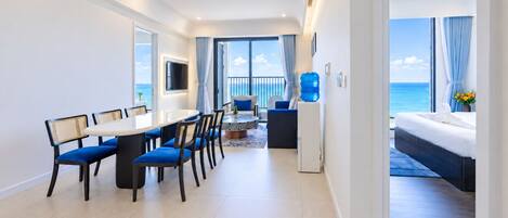 A modern hotel room with a dining area, a sofa, a large window with a view of the sea, and a bed with a view of the ocean.
