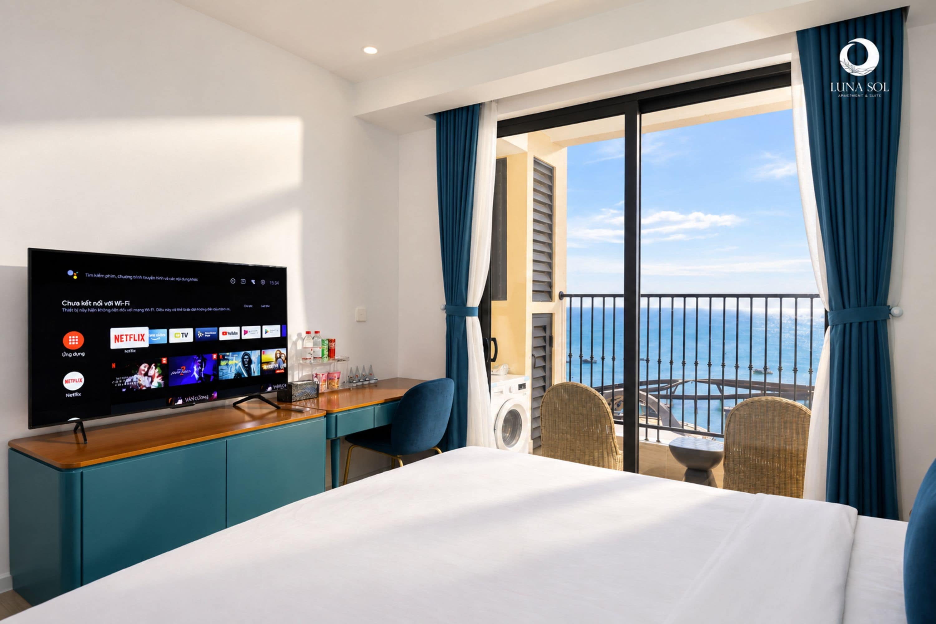 Een hotelkamer met een bed, een bureau met een computer, een televisie waarop streamingdiensten worden getoond, een balkon met uitzicht op zee, en een raam met gordijnen.