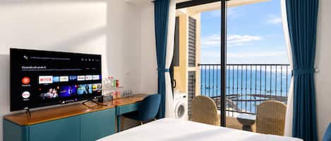 A hotel room with a bed, a desk with a computer, a TV displaying streaming services, a balcony with a view of the sea, and a window with curtains.