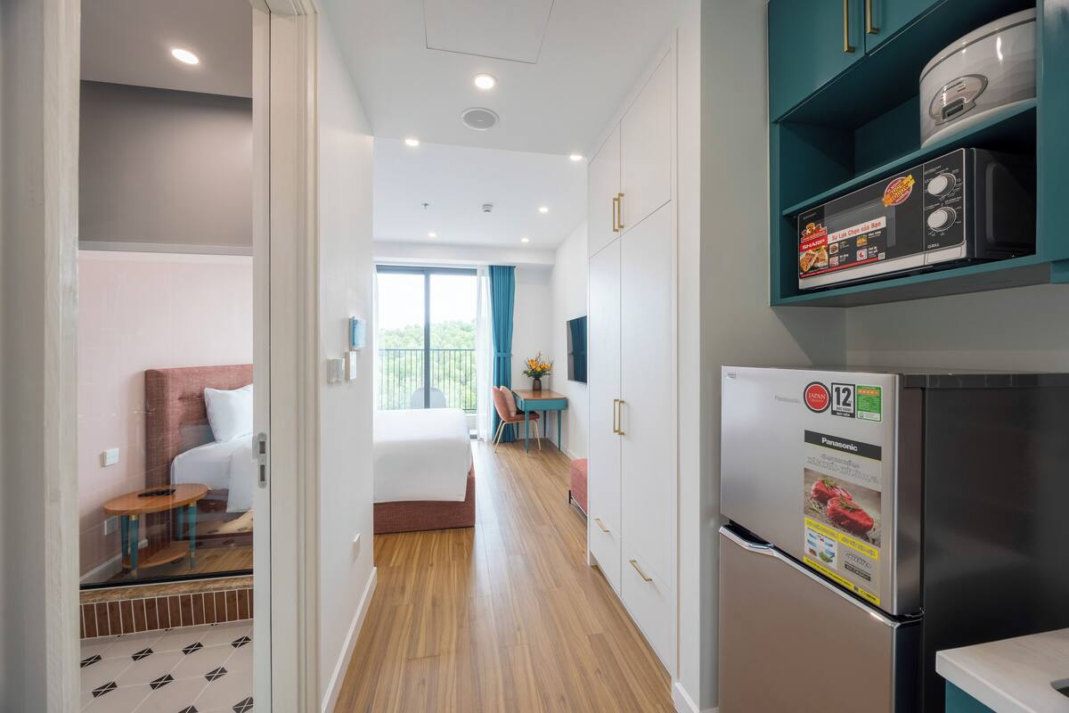 A modern hotel room with a bed, a small desk, a dining area, and a kitchen with a refrigerator and microwave.