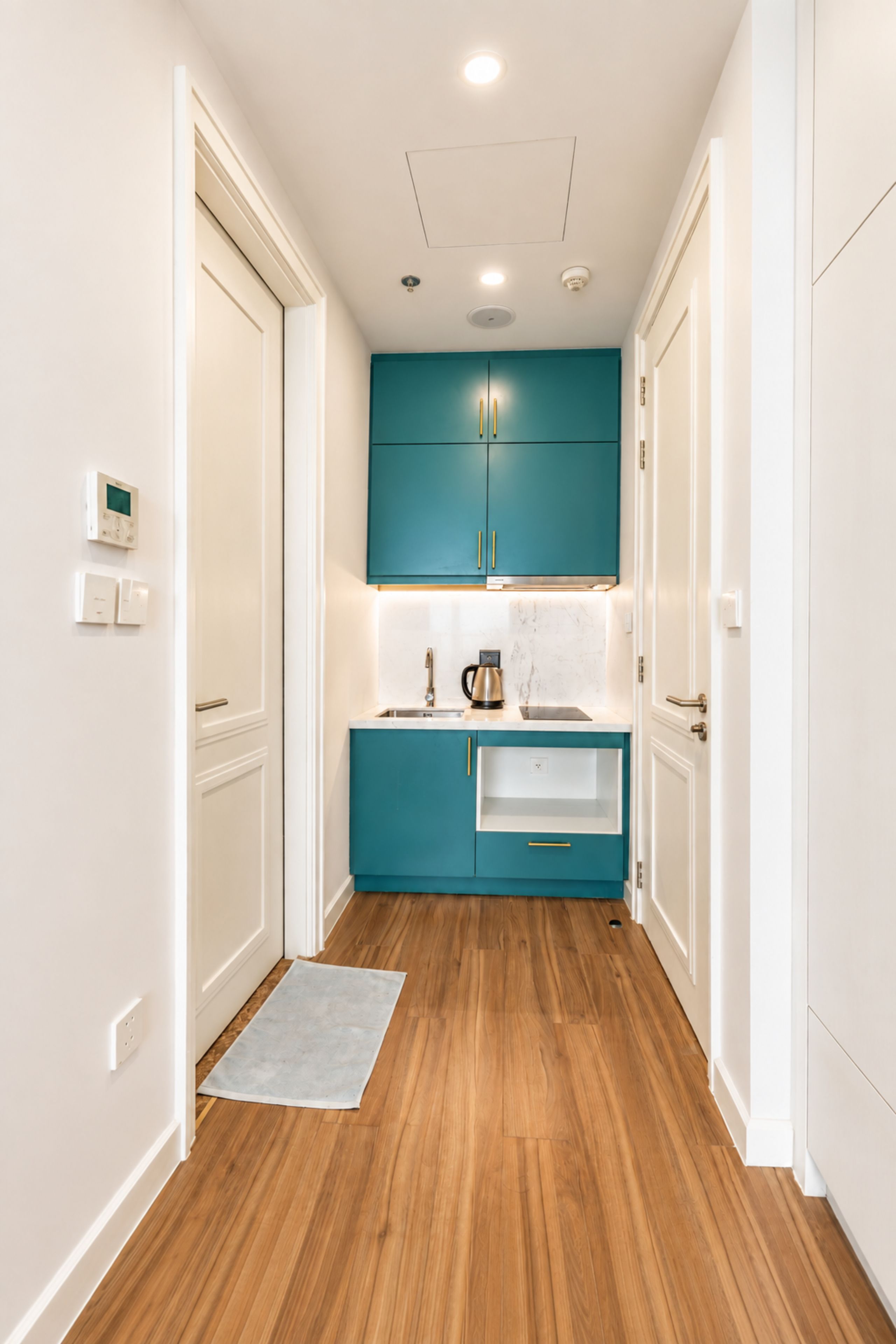 A narrow hallway with wooden flooring, white walls, and teal cabinets with gold handles.