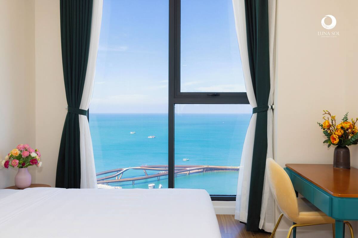 A hotel room with a large window offering a view of the sea and a bridge. There is a bed, a desk, and a chair.