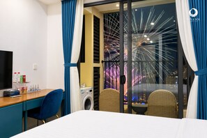 A hotel room with a bed, a desk with a chair, a washing machine, and a view of fireworks.