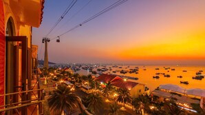 Vista costera al atardecer con un teleférico, embarcaciones y un edificio iluminado con palmeras.