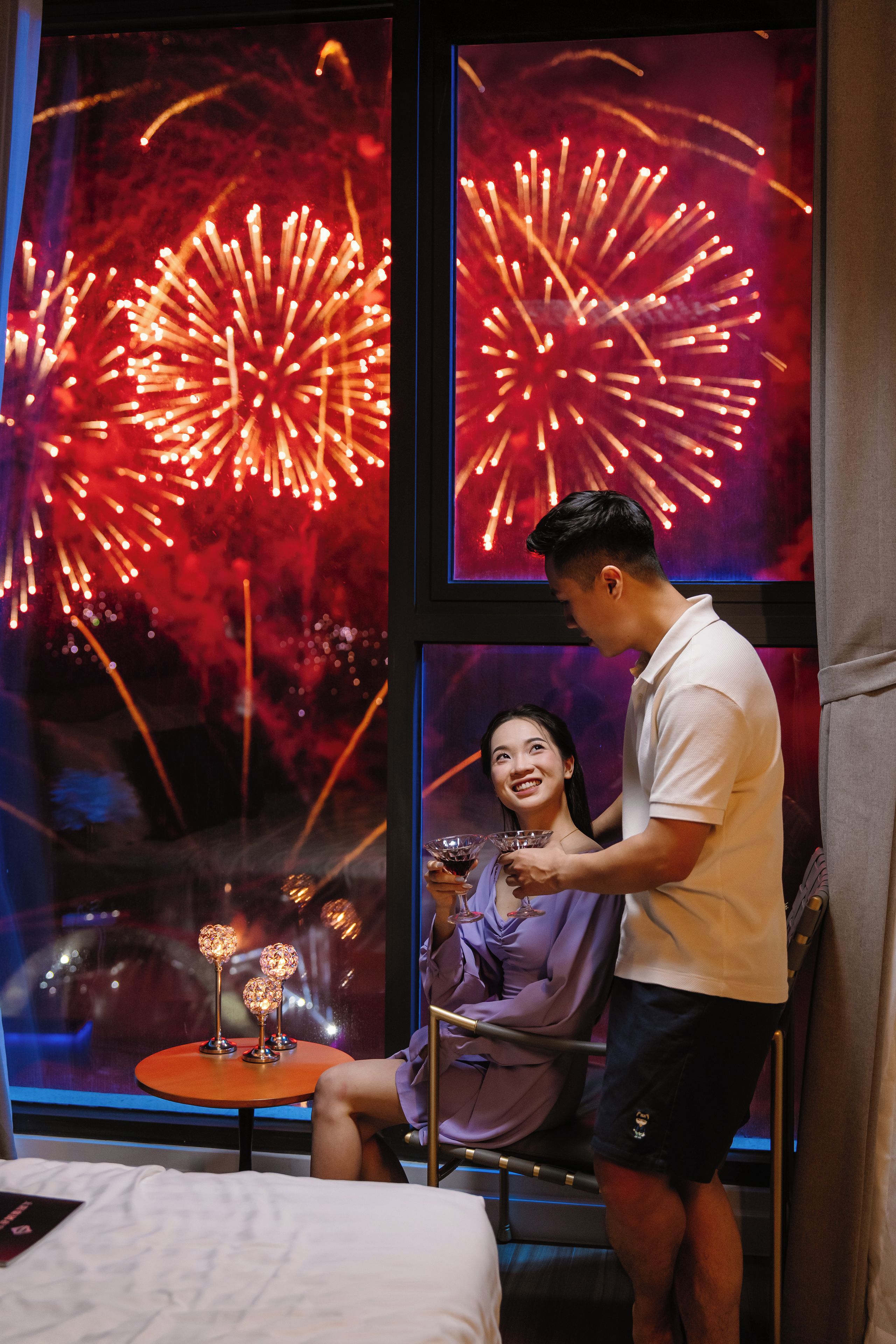 Une chambre dotée d’une grande fenêtre donnant sur un feu d’artifice, d’une petite table ronde agrémentée d’objets décoratifs et d’un lit recouvert d’une couette blanche.