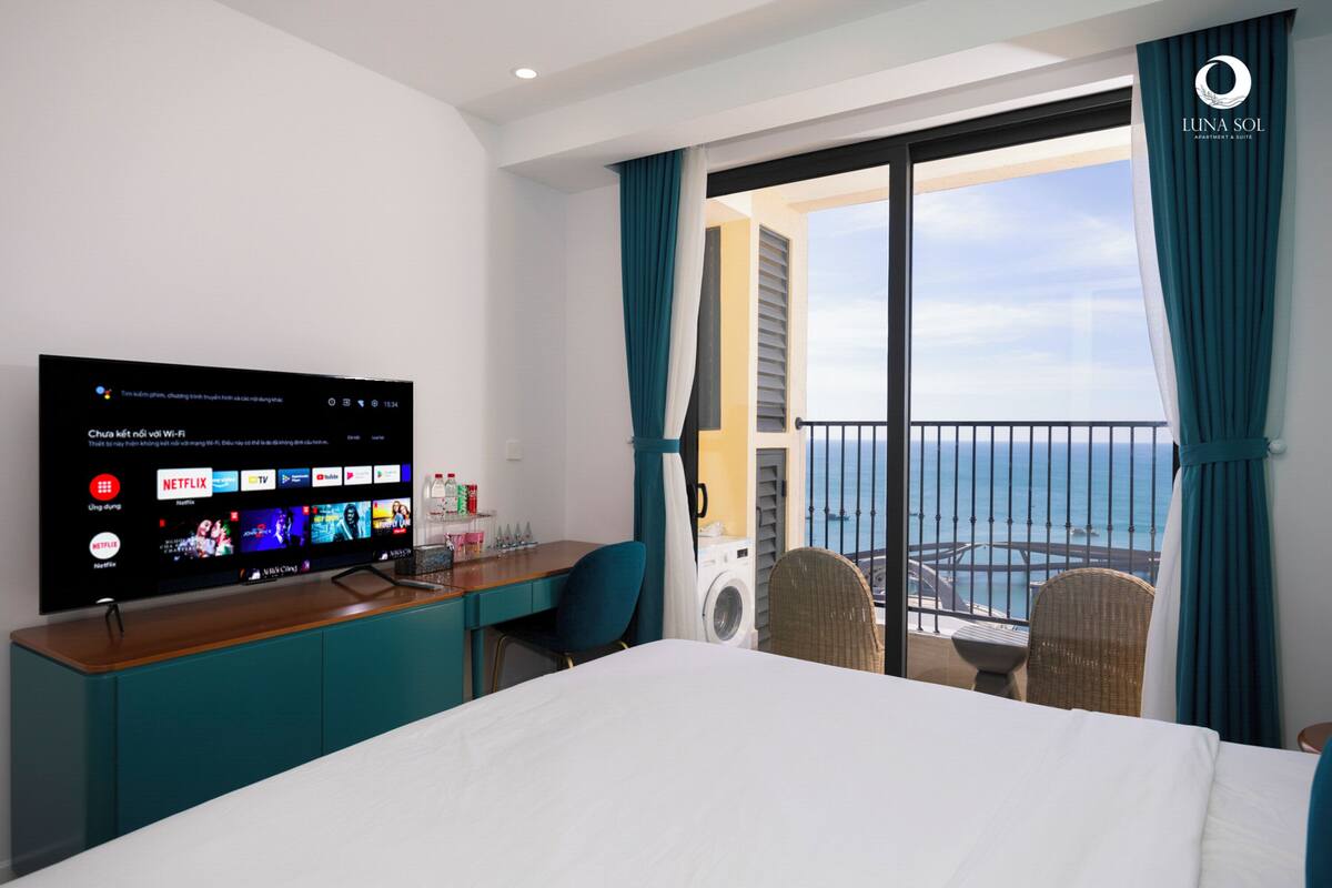 A hotel room with a large flat-screen TV displaying streaming services, a bed, a desk with a chair, a washing machine, and a balcony with a view of the sea.