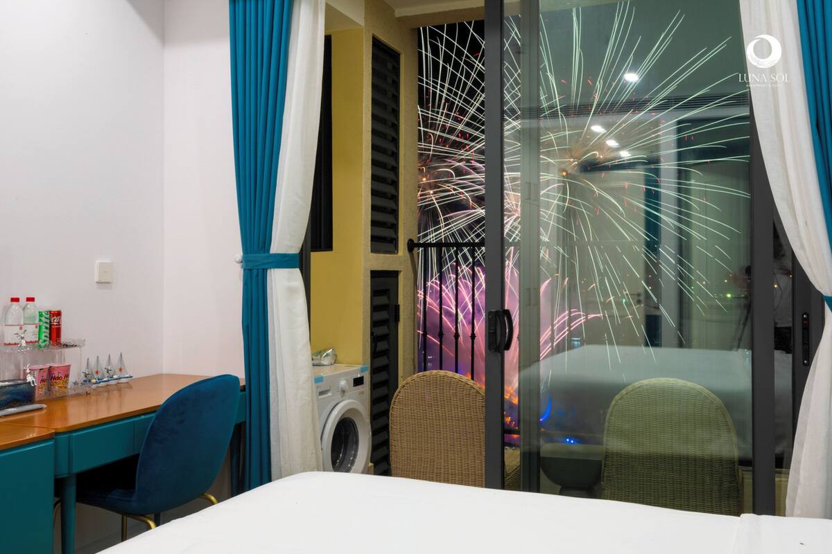 A hotel room with a bed, a desk, a chair, a washing machine, and a glass wall with a view of fireworks.