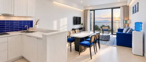 A modern kitchen with white cabinetry and blue tiles, a dining area with a table and chairs, and a balcony with a view of the sea.
