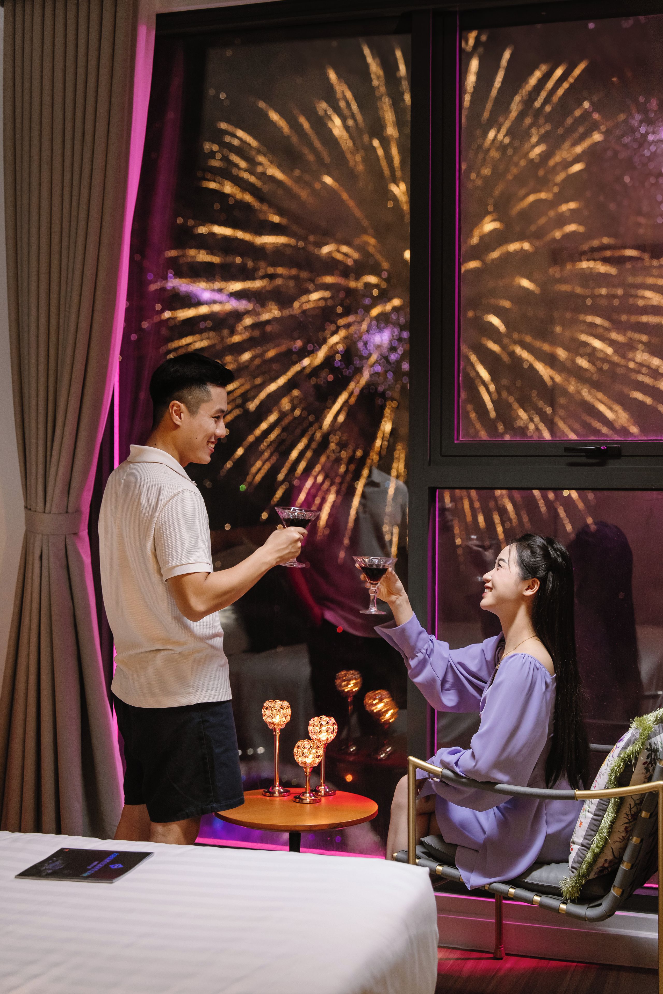 A man and a woman toasting with drinks in a room with a large window displaying fireworks.