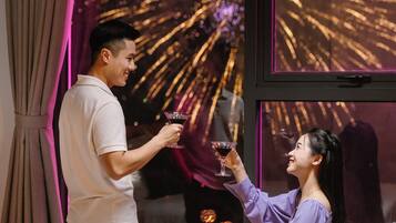 A man and a woman toasting with drinks in a room with a large window displaying fireworks.