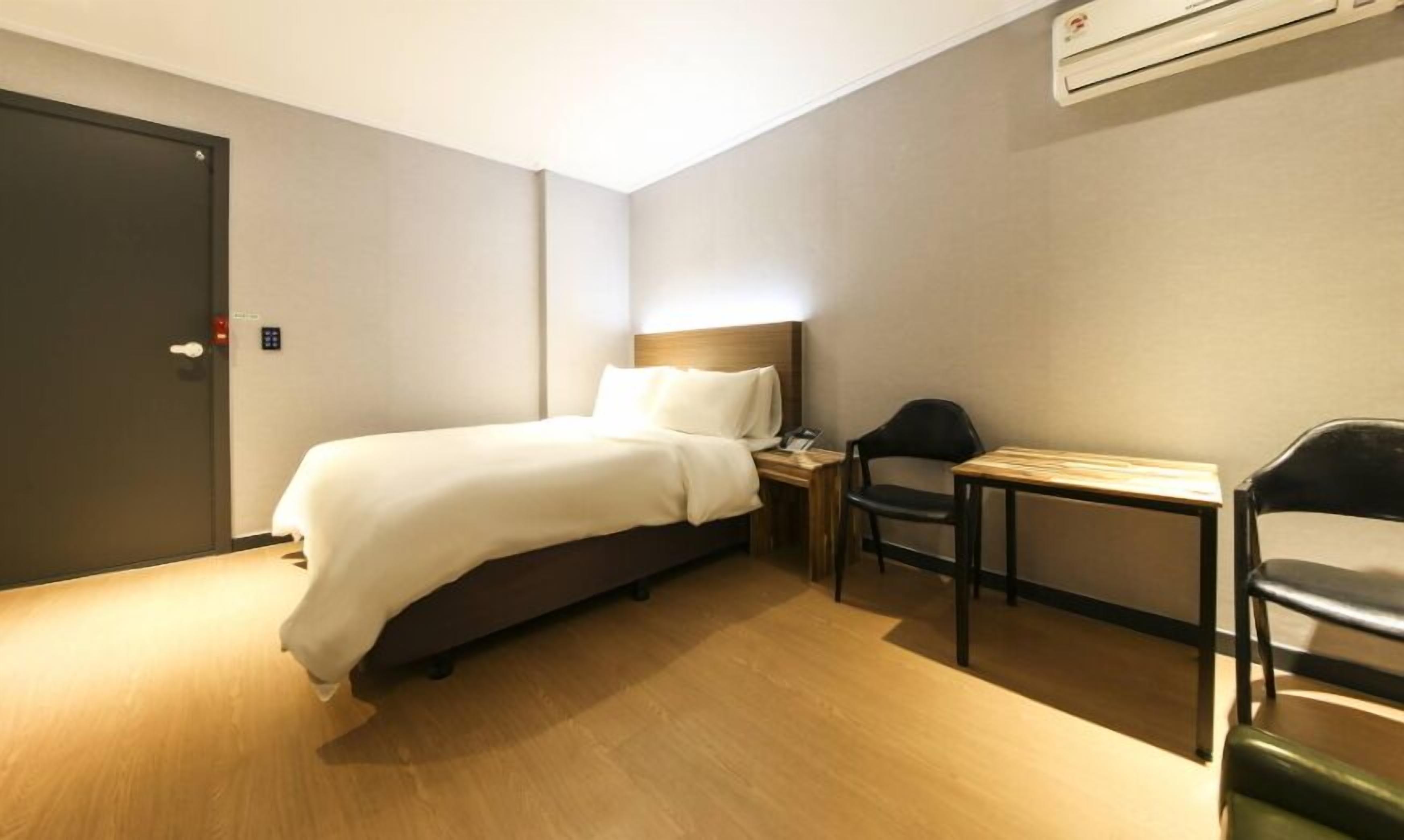 A compact hotel room with a single bed, a small desk, a chair, and a bedside table.