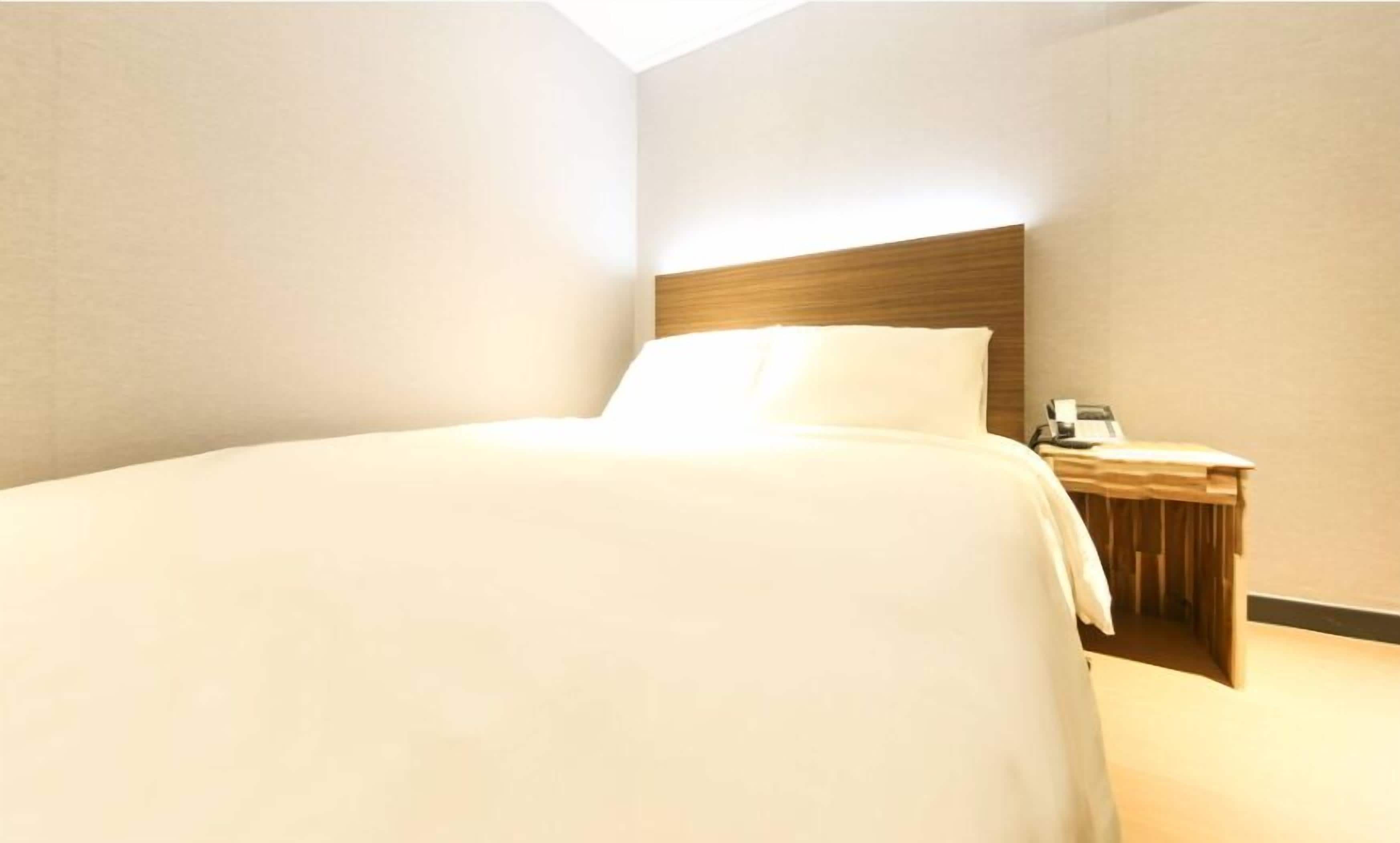 Una cama bien hecha con cabecera de madera en una habitación de paredes sencillas y una mesita de noche de madera a su lado.