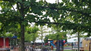 An outdoor seating area with tables and chairs, surrounded by trees and a playground in the background. - Tinyhouse / chalet at the Rhederlaagse Meren vacation park in NL-Lathum by the lake (Lathum)