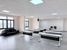 Un aula con mesas blancas, un sofá negro y ventanas amplias.