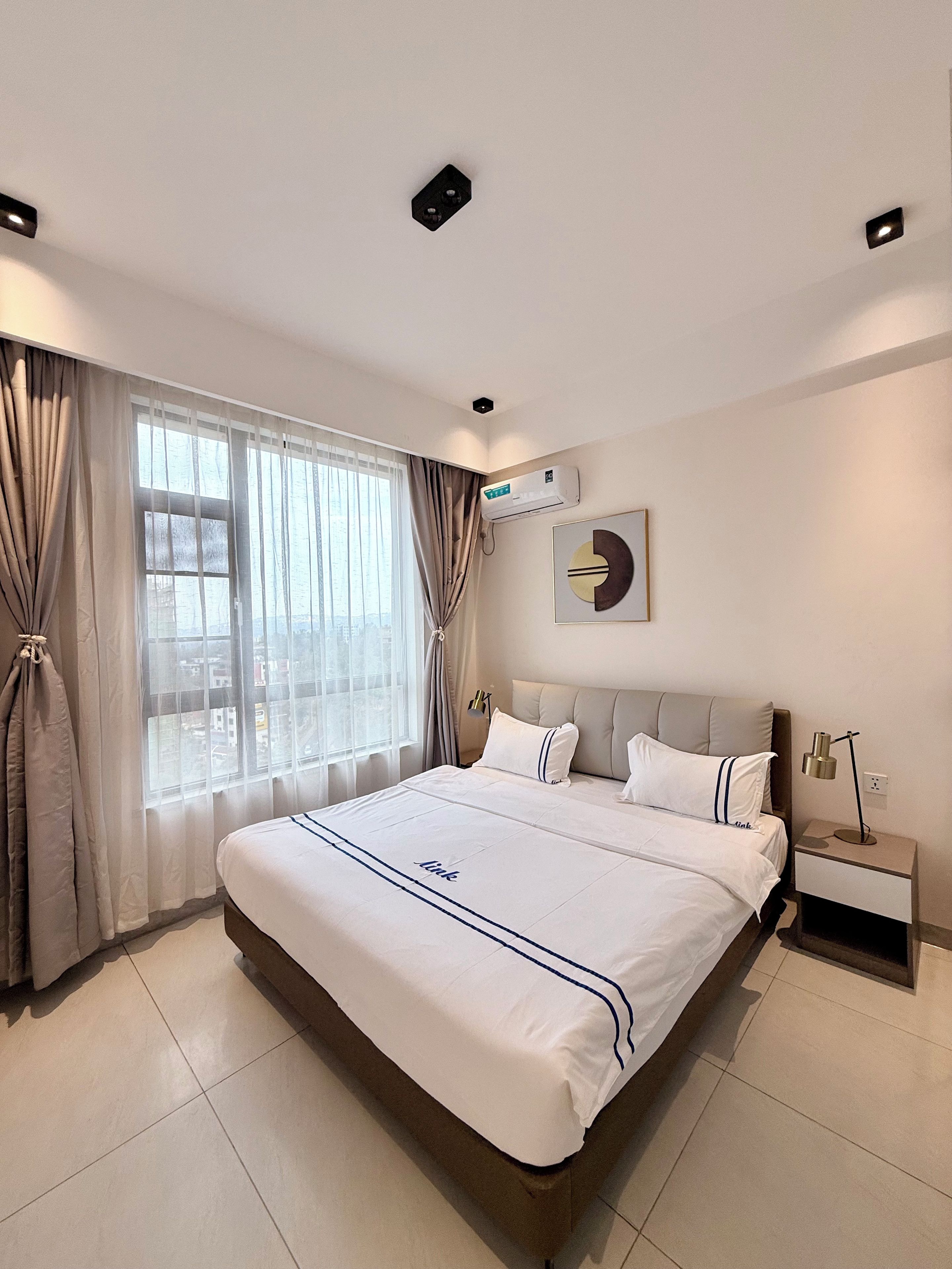A hotel room with a large bed, a bedside table with a lamp, an air conditioning unit, and a view through the window.