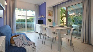 A modern living room with a sofa, a dining table with chairs, a TV, and a large sliding glass door leading to an outdoor area.