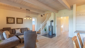 A spacious living area with a sofa, armchair, and wooden flooring. A staircase leads to an upper level, and a fireplace is visible in the background.