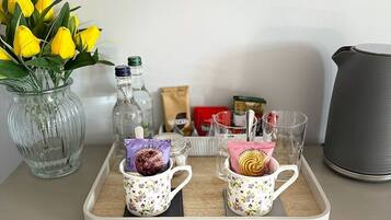 Una bandeja con dos tazas florales que contienen alimentos empaquetados, un jarrón con tulipanes amarillos y una tetera sobre una mesa.
