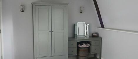 A bedroom with a grey wardrobe, a bedside table, a mirror, and a waste bin.