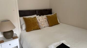 A neatly made bed with white bedding, a dark headboard, and three pillows. A lamp and a small drawer are visible on the nightstand beside the bed.