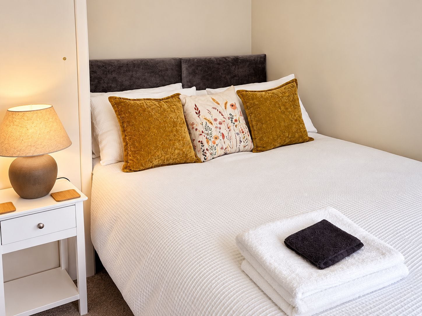 A neatly made bed with white bedding, two mustard-colored pillows, and a floral-patterned pillow. A lamp is on a bedside table, and a folded towel is on the bed.