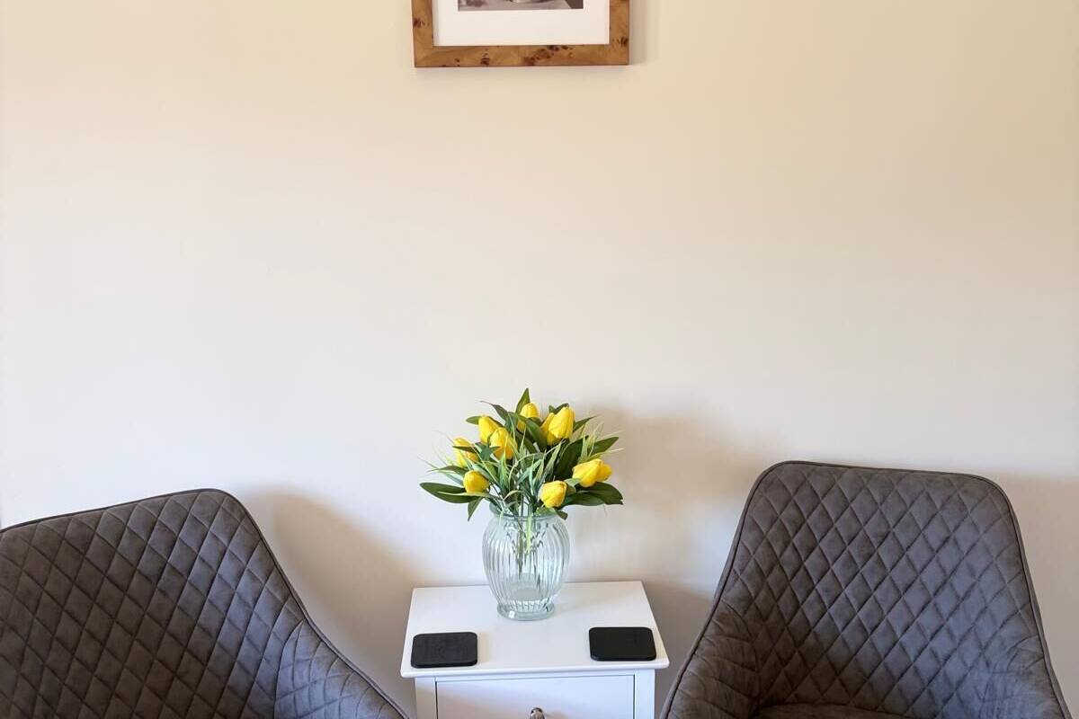 Two grey upholstered chairs, a small white nightstand with a vase of yellow flowers, and a framed picture on the wall.