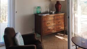 A room with a wooden ceiling, a brown armchair, a wooden cabinet with drawers, a glass door leading outside, and a small table with a lamp. - Mondomini Panoramic Cottage, stunning view on sea, hills and 8500 olive trees (Marche)