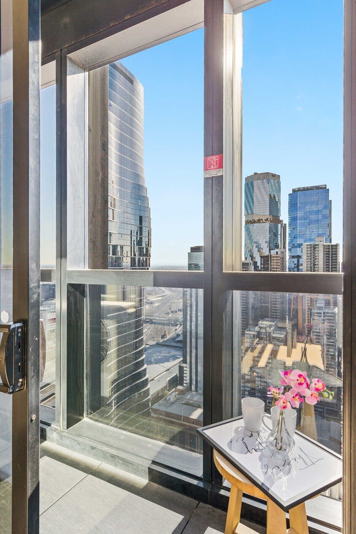 A modern hotel room with a large window offering a city view, a small table with a vase of flowers, and a door with a security lock.