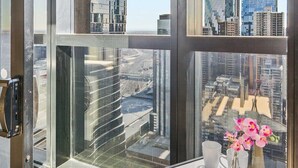 Quarto de hotel moderno com uma grande janela com vista para a cidade, uma mesa pequena com um vaso de flores e uma porta com tranca de segurança.