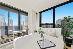 A modern hotel room with a large bed, a desk, and a view of the city skyline through large windows. - Modern 2BR on Collins, Skyline Views & Parking (Melbourne)
