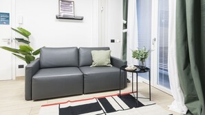 Una sala moderna con un sofá gris, una mesita auxiliar con un jarrón y una taza, una planta verde grande y un tapete blanco y negro.