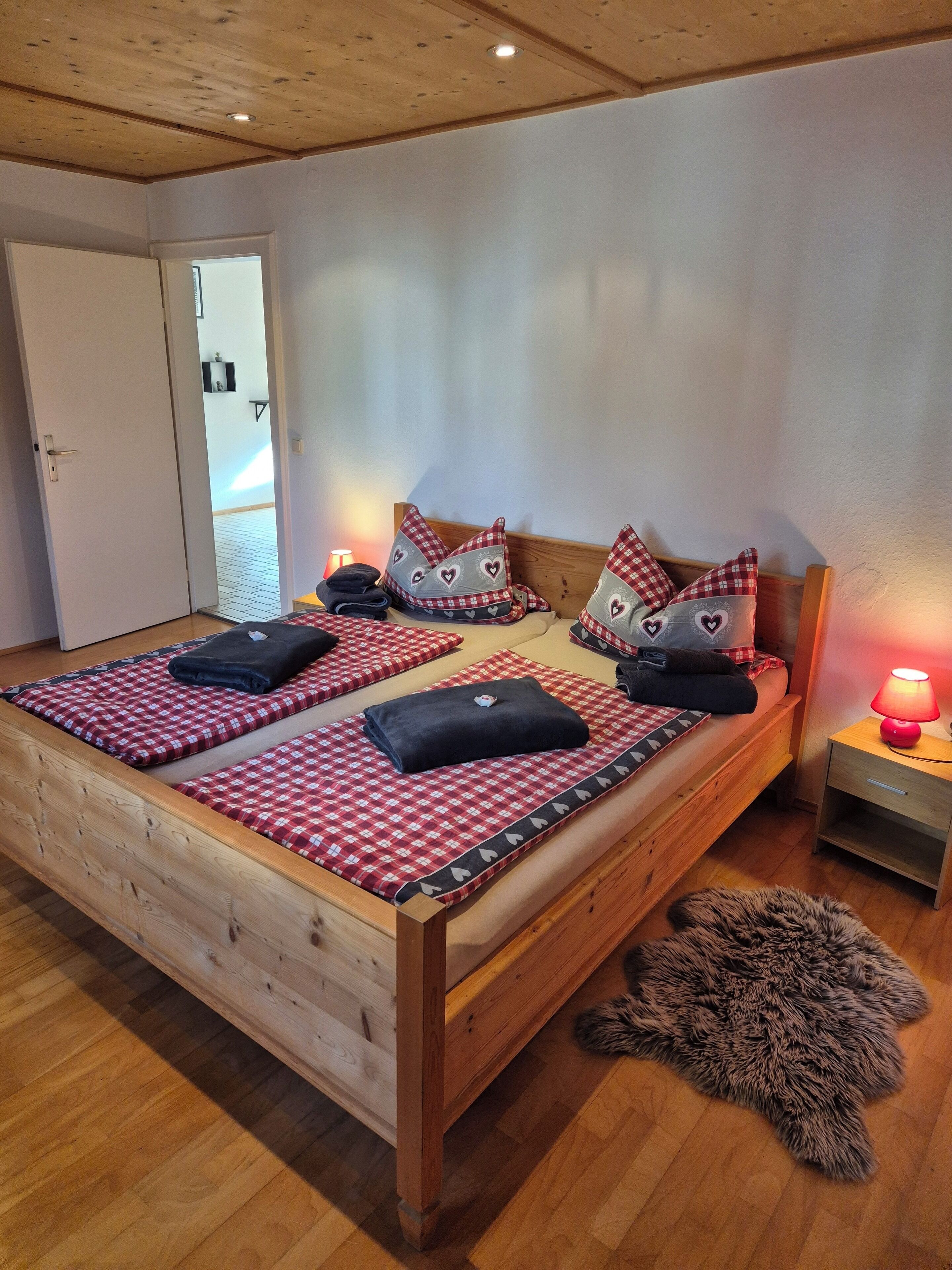 A wooden bed with checkered bedding and heart-patterned pillows, a small bedside table with a lamp, and a fur rug on the floor.