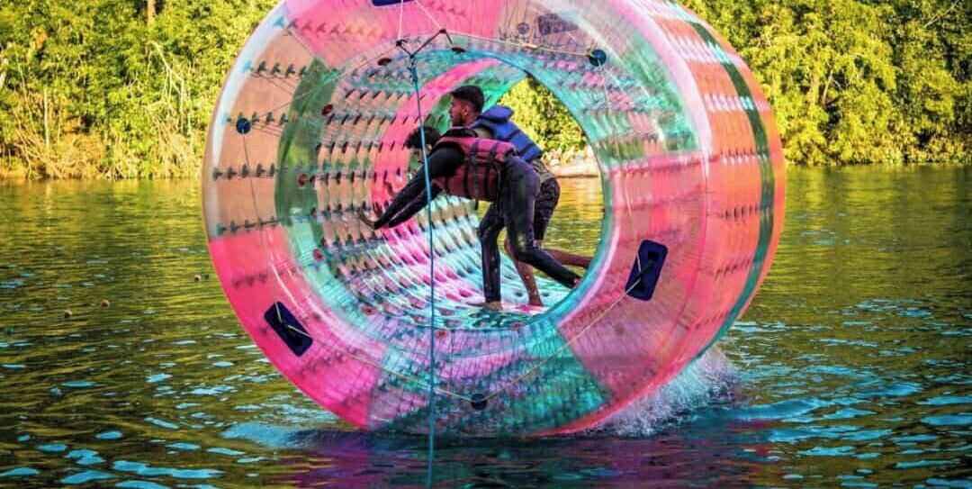 A person inside a transparent, cylindrical water tube with a person outside, both wearing life jackets, floating on water.