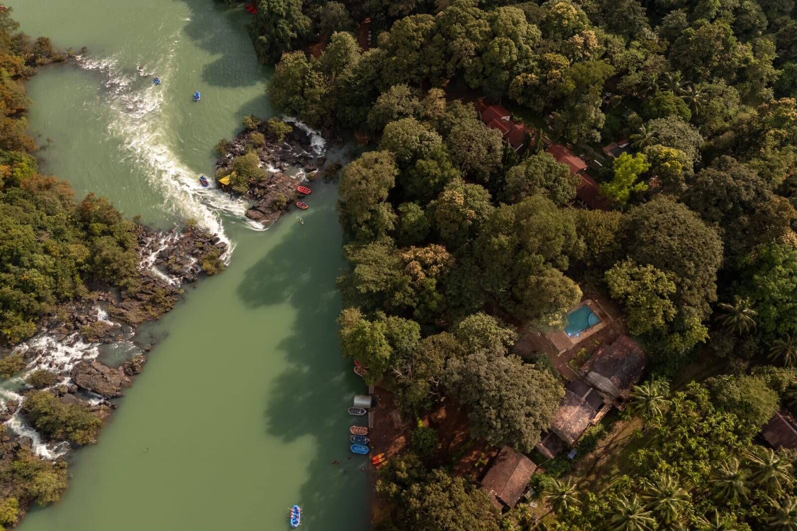 An aerial view of a riverside property with a swimming pool, surrounded by dense forest and rocky terrain.