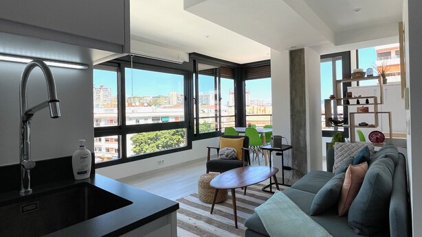 Un apartamento moderno con cocina, comedor y sala de estar. Cuenta con ventanas amplias que ofrecen vistas de la ciudad, un sofá, una mesa de comedor y un lavamanos de cocina.