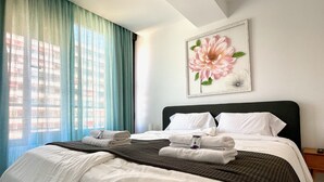 Una cama bien hecha con sábanas blancas y un cabecero oscuro, una ventana con cortinas azules, y una obra de arte floral enmarcada en la pared.