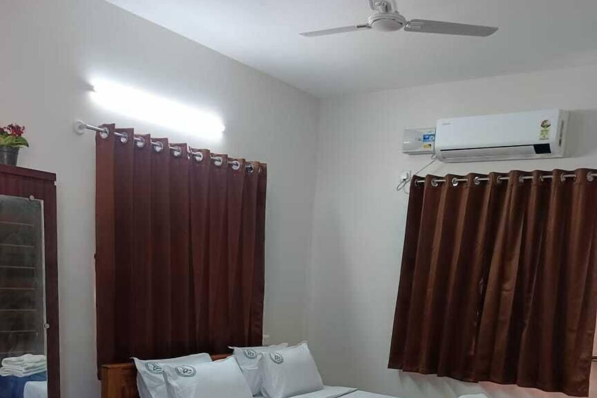A bedroom with a wooden bed, white bedding, a blue bed runner, a wooden nightstand, a mirror, a ceiling fan, and an air conditioning unit.