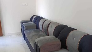 A long, dark-colored sofa with multiple cushions against a plain wall, a tiled floor, and a power outlet on the wall. - Rithu's inn (Tirupattur)