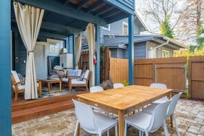 Een terras buiten met een houten tafel en witte stoelen, een overdekt zitgedeelte met kussens, en een houten omheining.