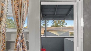 Un espace extérieur abrité avec une chaise rouge, une fenêtre ornée de rideaux à motifs et une vue sur un bâtiment doté d’un toit en pente.