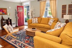 A living room with a sofa, armchair, and a coffee table. There is a door, a plant, and a wall with a picture frame. - Grand Boho - Team Joseph Ellen (Jacksonville)