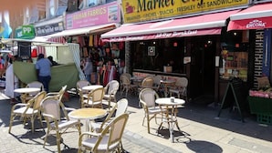 Outdoor seating with wicker chairs and tables, a Market Cafe sign, and a storefront with a sign for RAIHAN FABRICS. - Walthamstow - Apartment - Stay 3 - Street Parking (London)