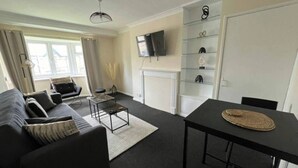 A modern living room with a sofa, coffee table, and dining table. There is a fireplace, a TV mounted on the wall, and a bookshelf. - Walthamstow - Apartment - Stay 3 - Street Parking (London)
