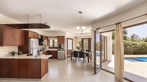 A modern kitchen with a central island, a dining area with a table and chairs, and a view of a swimming pool through large sliding glass doors. - Villa Tsikkos Pende in Argaka (Argaka)