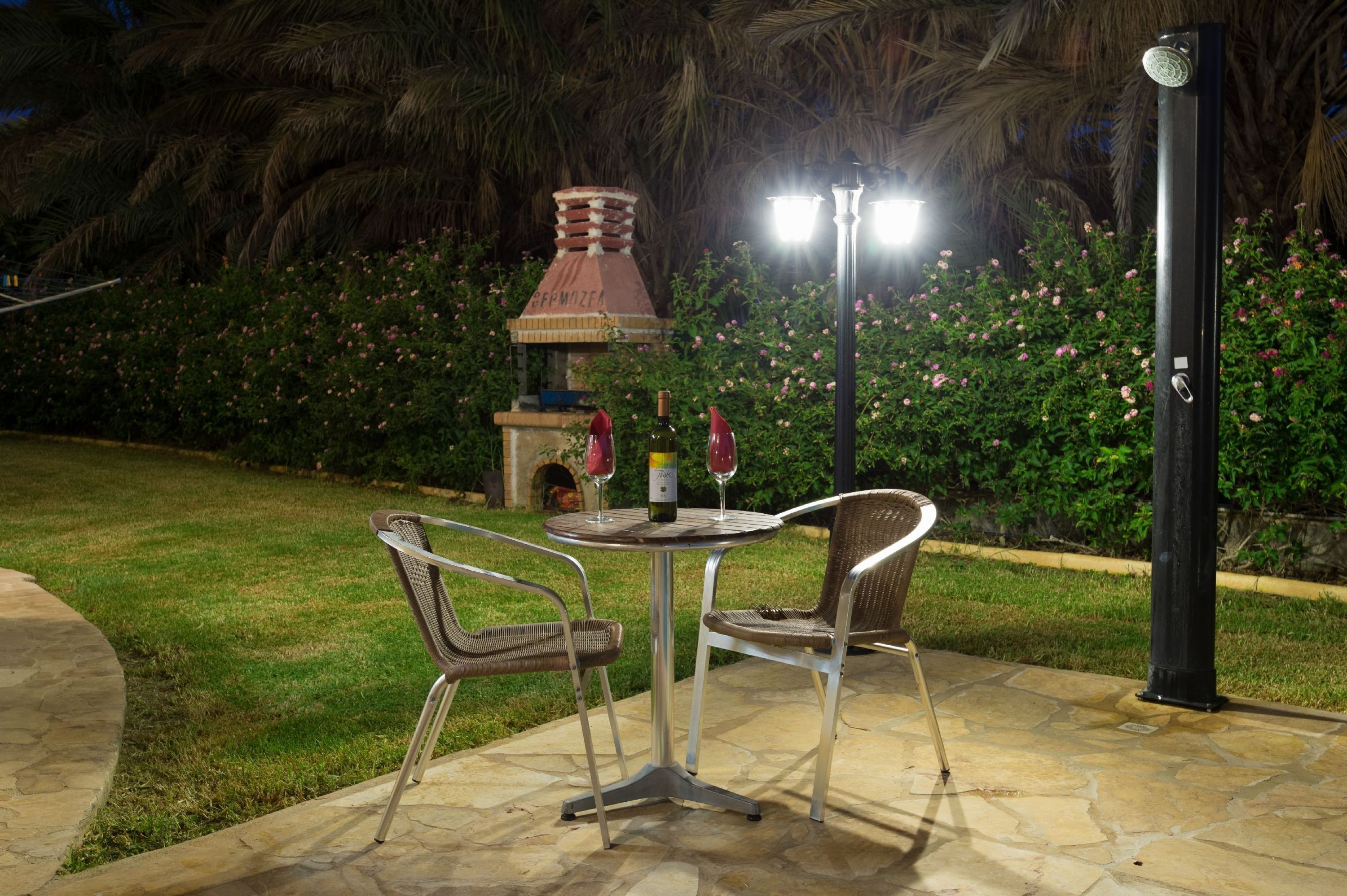 Une terrasse extérieure avec une table ronde et deux chaises, une cheminée en briques et un espace bien éclairé avec un lampadaire noir.